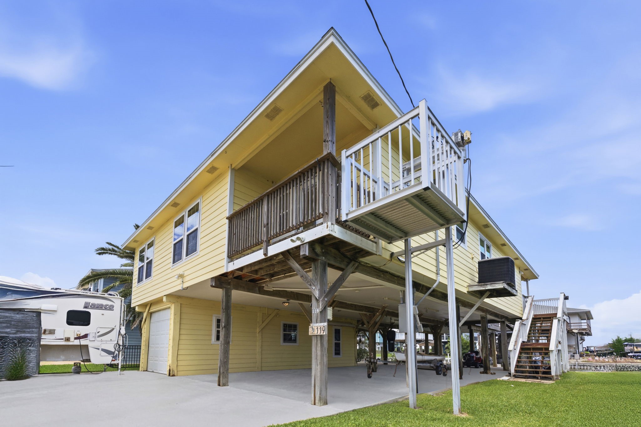 119 Sand Shoals Road Freeport, TX 77541 - Photo 32 of 34 Front exterior — elevated yellow cement-board home on pilings with covered upper porch, white railings, ground-level garage, covered carport, and dedicated boat/trailer parking.