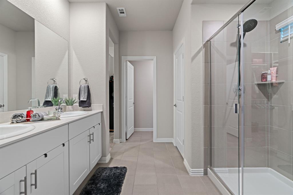 3112 Kingsgarden Road Denton, TX 76207 - Photo 30 of 38 Bathroom featuring a double vanity with white cabinetry and undermount sinks, a large frameless mirror, and a glass-enclosed shower with tiled walls and a built-in shelf
