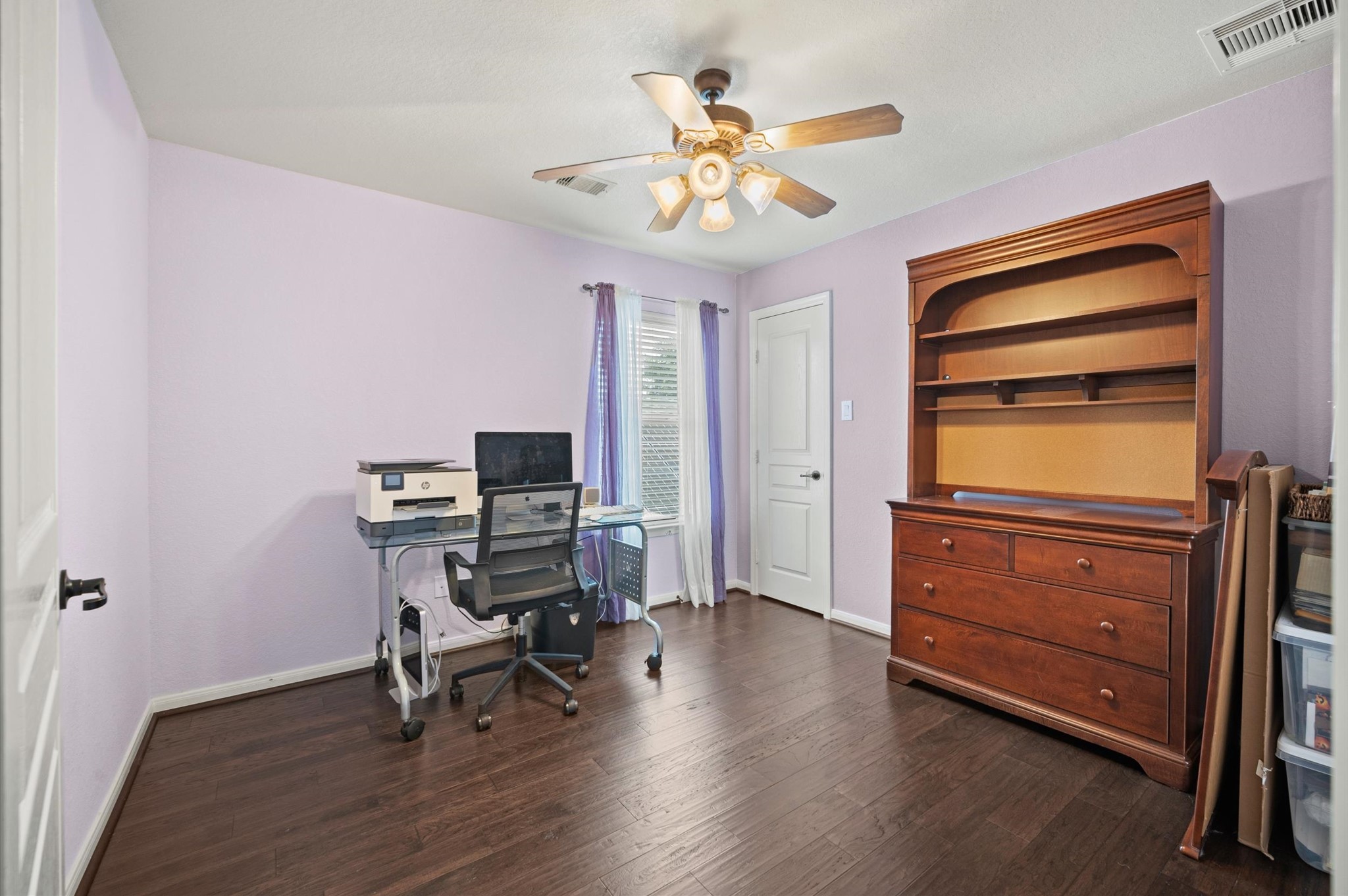 9507 Empress Crossing Drive Spring, TX 77379 - Photo 35 of 50 This room is a bright and functional space with light purple walls and dark hardwood floors. It features a ceiling fan, a window with natural light, a glass desk with office equipment, and a wooden hutch with storage. Ideal for a home office or study.