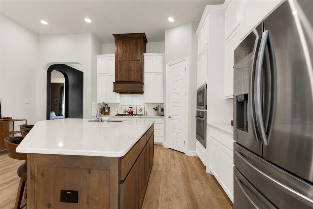 6404 Trezevant Street Rowlett, TX 75089 - Photo 6 of 30 Spacious kitchen featuring a large central island with a light-toned countertop, wood-finish base cabinetry, a stainless steel sink with a gooseneck faucet, a substantial wood-paneled range hood, and white upper and lower cabinetry