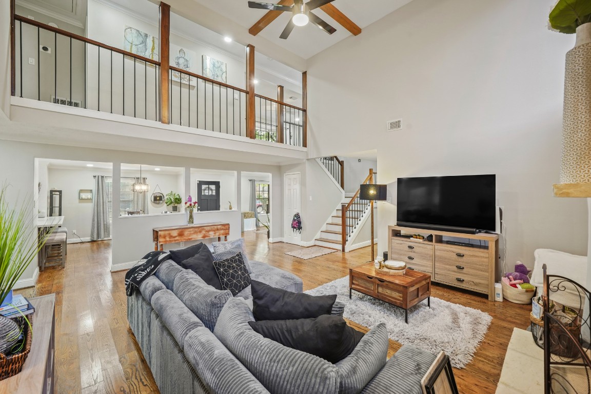 This spacious living room features high ceilings, hardwood floors, and an open layout. A large TV sits on a modern entertainment unit, and a plush sectional sofa provides ample seating. The room is bright and airy with a view of the upstairs loft area, accessible by a stylish staircase.