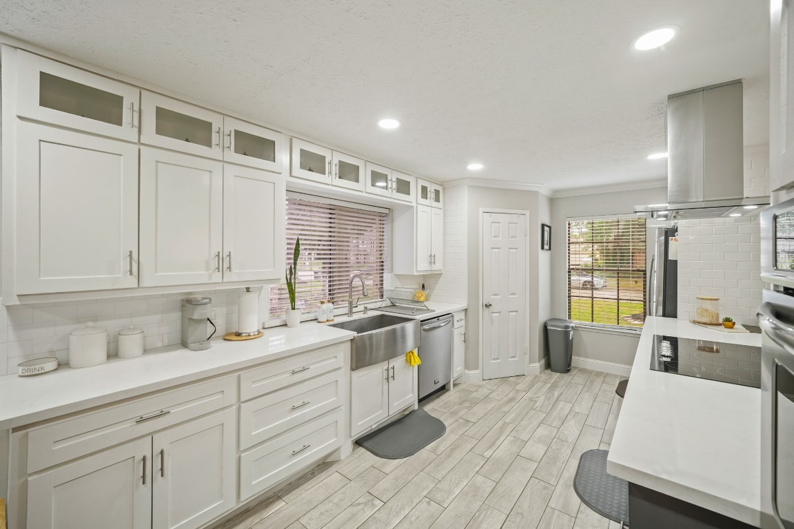 15831 Ridge Park Drive Houston, TX 77095 - Photo 4 of 26 This photo showcases a modern kitchen with sleek white cabinetry, stainless steel appliances, and wood-look tile flooring. It features ample counter space, a farmhouse sink, and large windows for natural light. The layout is open and functional, perfect for cooking and entertaining.