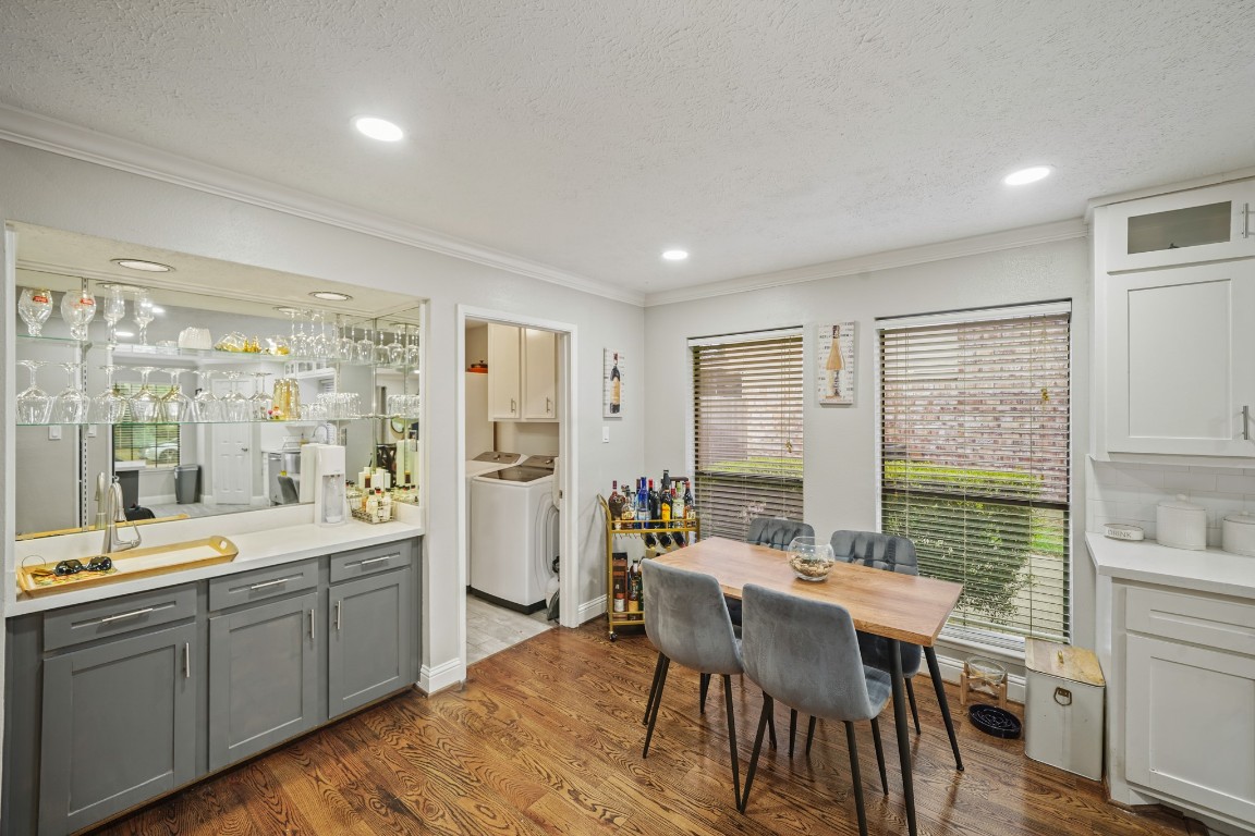 15831 Ridge Park Drive Houston, TX 77095 - Photo 7 of 26 This photo showcases a cozy dining area with a wooden table and four chairs, adjacent to a bar with glass shelving and cabinetry. The room features hardwood floors, large windows with blinds, and recessed lighting, providing a bright and inviting space. A doorway leads to a utility room with a washer and dryer.