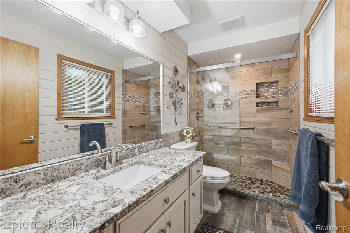 1221 Rhodes Road Lake Orion, MI 48360 - Photo 34 of 72 Bathroom featuring a single vanity with a white countertop and undermount sink, a walk-in shower with natural stone-look tile, a glass shower enclosure, and wood-finish flooring