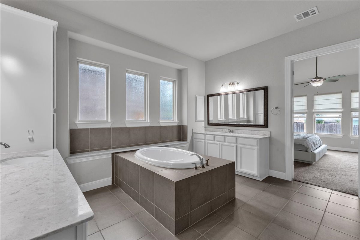 21301 Resource Road Pflugerville, TX 78660 - Photo 12 of 29 Bathroom featuring a built-in soaking tub with tile surround, two separate vanities with stone countertops, and three privacy windows