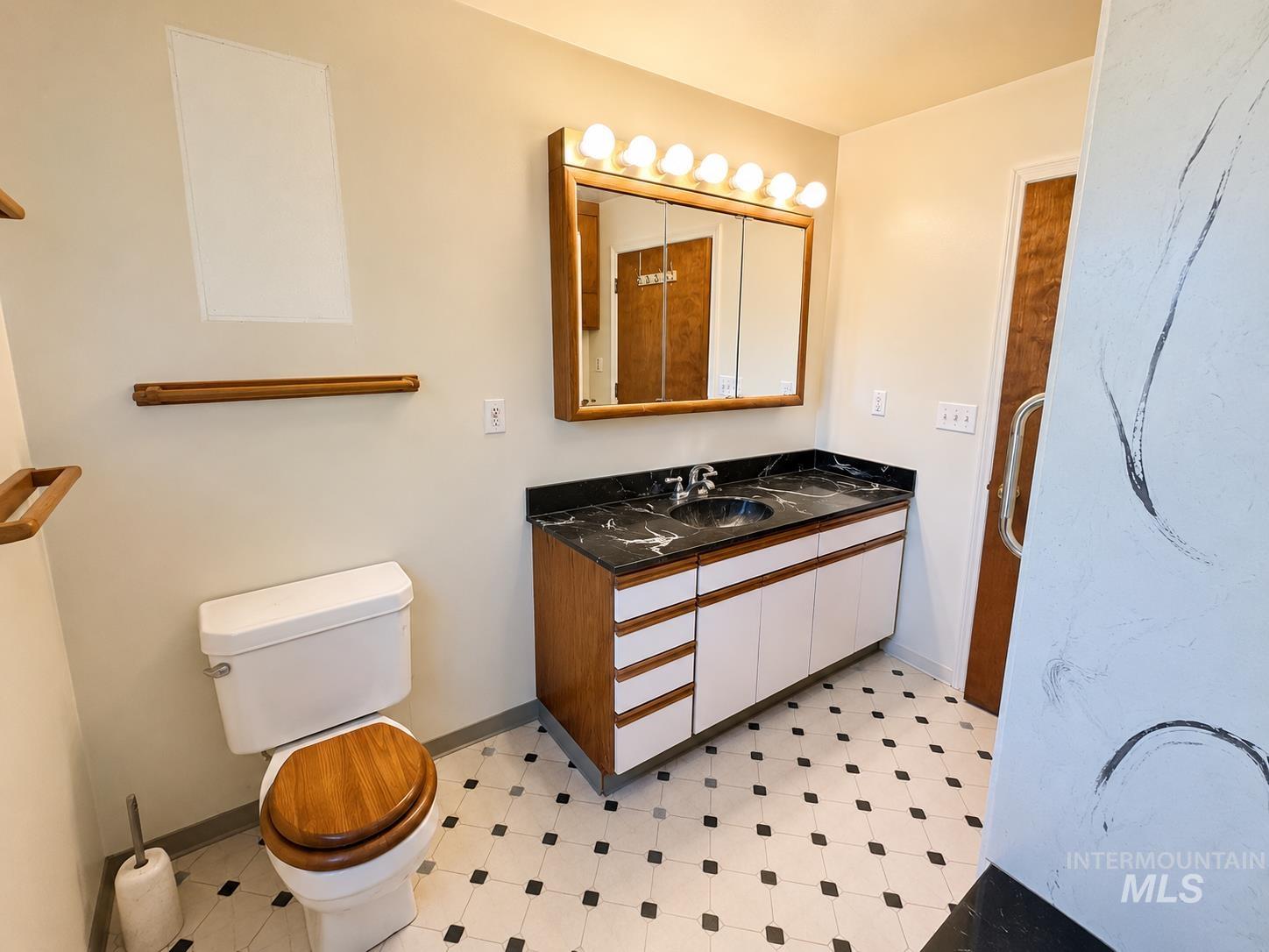 16434 Pride Lane Caldwell, ID 83607 - Photo 13 of 16 Bathroom featuring a vanity with a black countertop and integrated sink, a large mirrored medicine cabinet with overhead lighting, a toilet with a wood-finish seat, a wood-finish towel bar, and patterned floor tiling