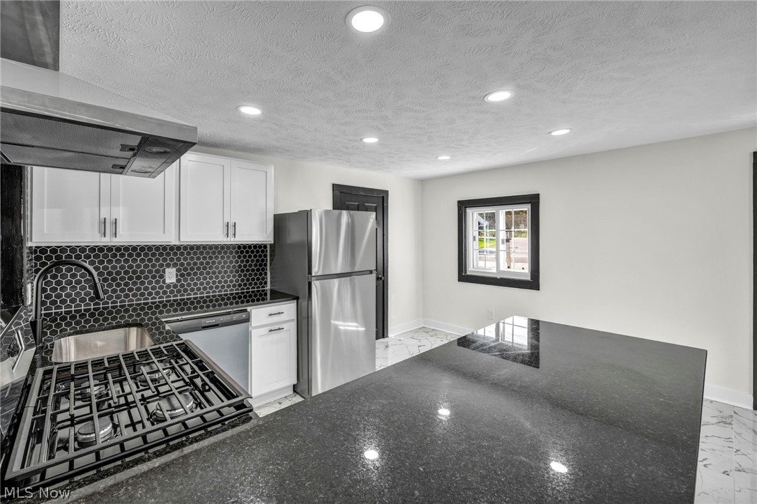 7339 River Road Olmsted Falls, OH 44138 - Photo 12 of 45 Modern kitchen featuring a black granite-style island, white cabinetry with black hardware, black hexagonal tile backsplash, stainless steel appliances, and recessed lighting