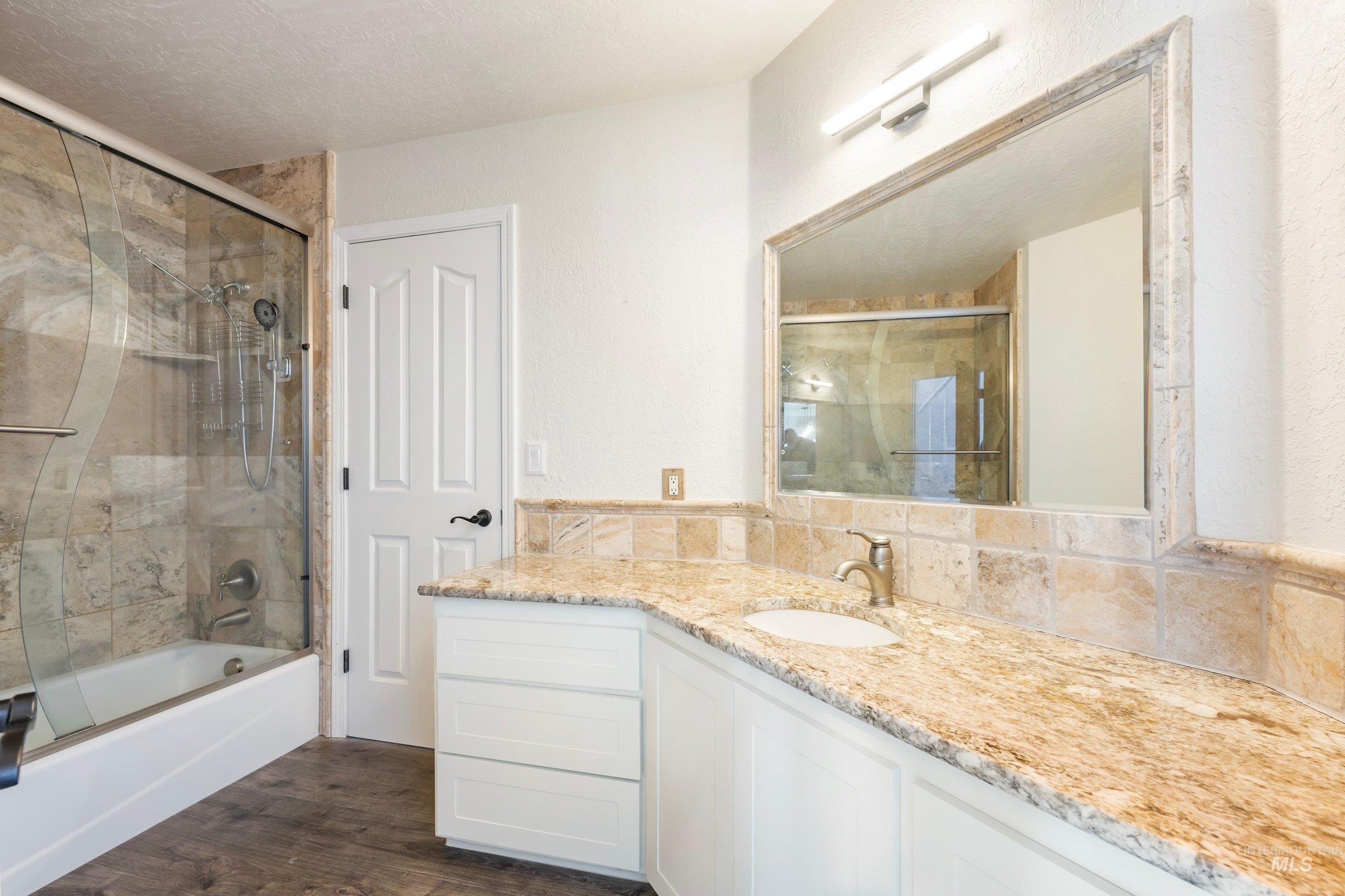 7657 Track Road Nampa, ID 83686 - Photo 18 of 49 Bathroom featuring a stone-tiled tub/shower combination with a clear glass enclosure, a white vanity with panel-front cabinetry, a light-toned stone countertop, a rectangular undermount sink, and wood-finish flooring