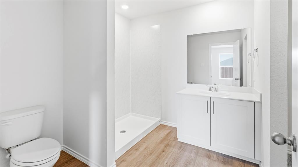 3319 Twilight Trail Forney, TX 75126 - Photo 19 of 33 Bathroom featuring wood-finish flooring, a white single-sink vanity with cabinet storage, a frameless mirror, a white toilet, and a stand-up shower with textured wall panels