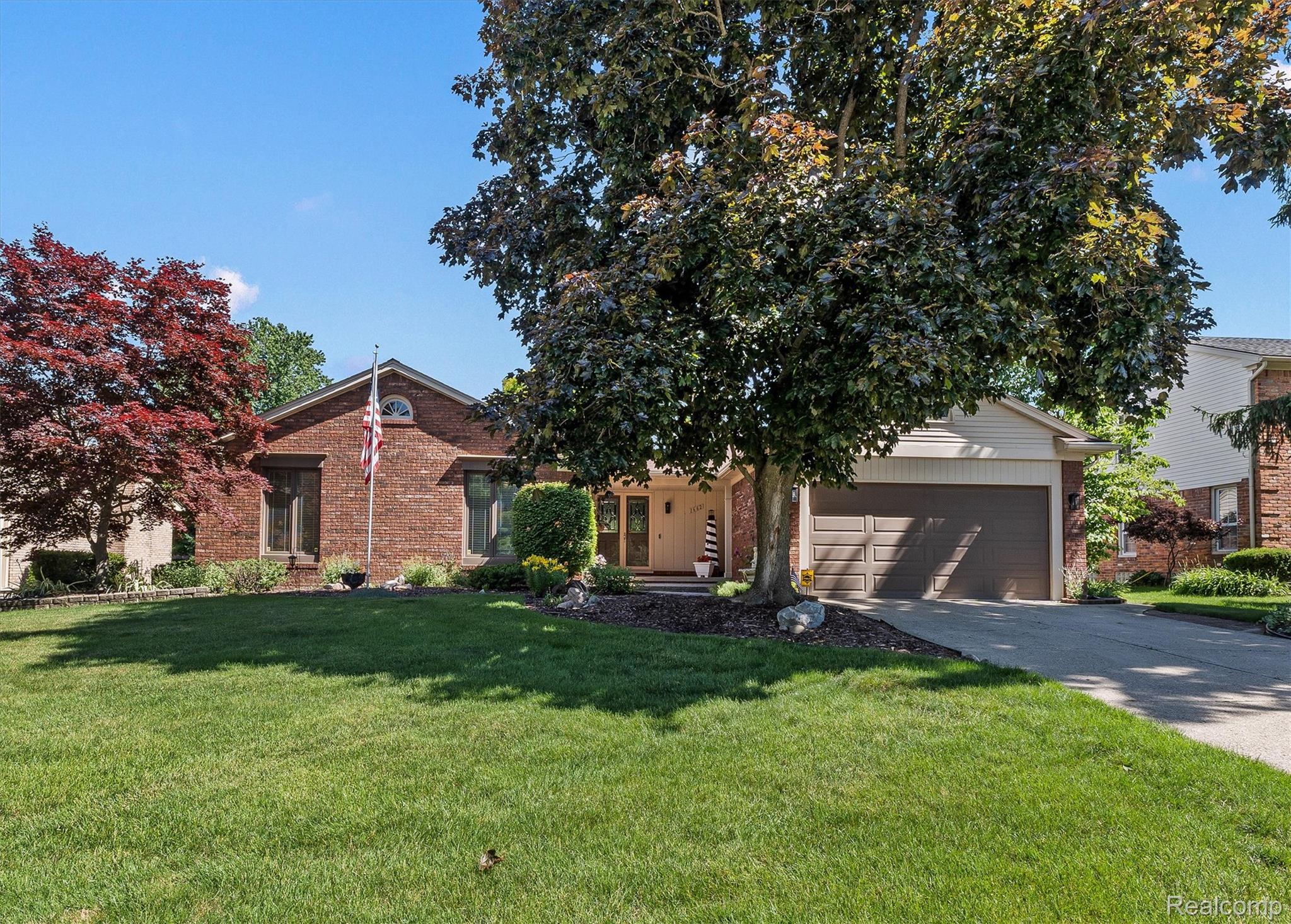 Brick exterior featuring a two-car attached garage, expansive front lawn, mature trees, and a concrete driveway