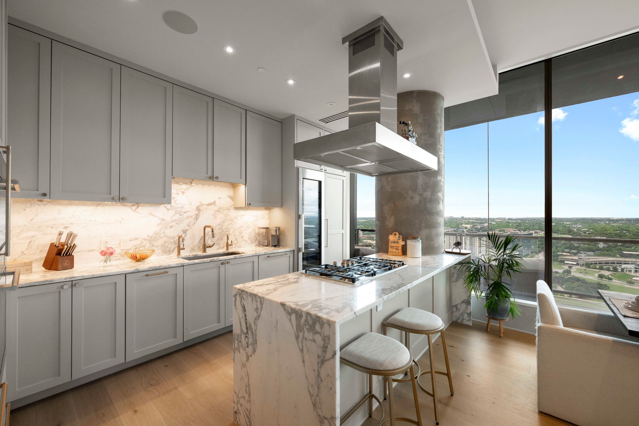 210 Lavaca Street, Unit 2706 Austin, TX 78701 - Photo 5 of 32 Contemporary kitchen featuring a waterfall island, integrated cooktop with suspended range hood, full-height windows, light wood-finish flooring, and light gray cabinetry with under-cabinet lighting