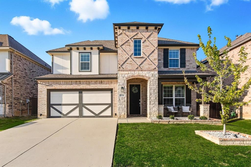 Two-story residence featuring a brick and stone facade, covered front porch, an attached two-car garage with decorative doors, a concrete driveway, and landscaped front yard with a tree