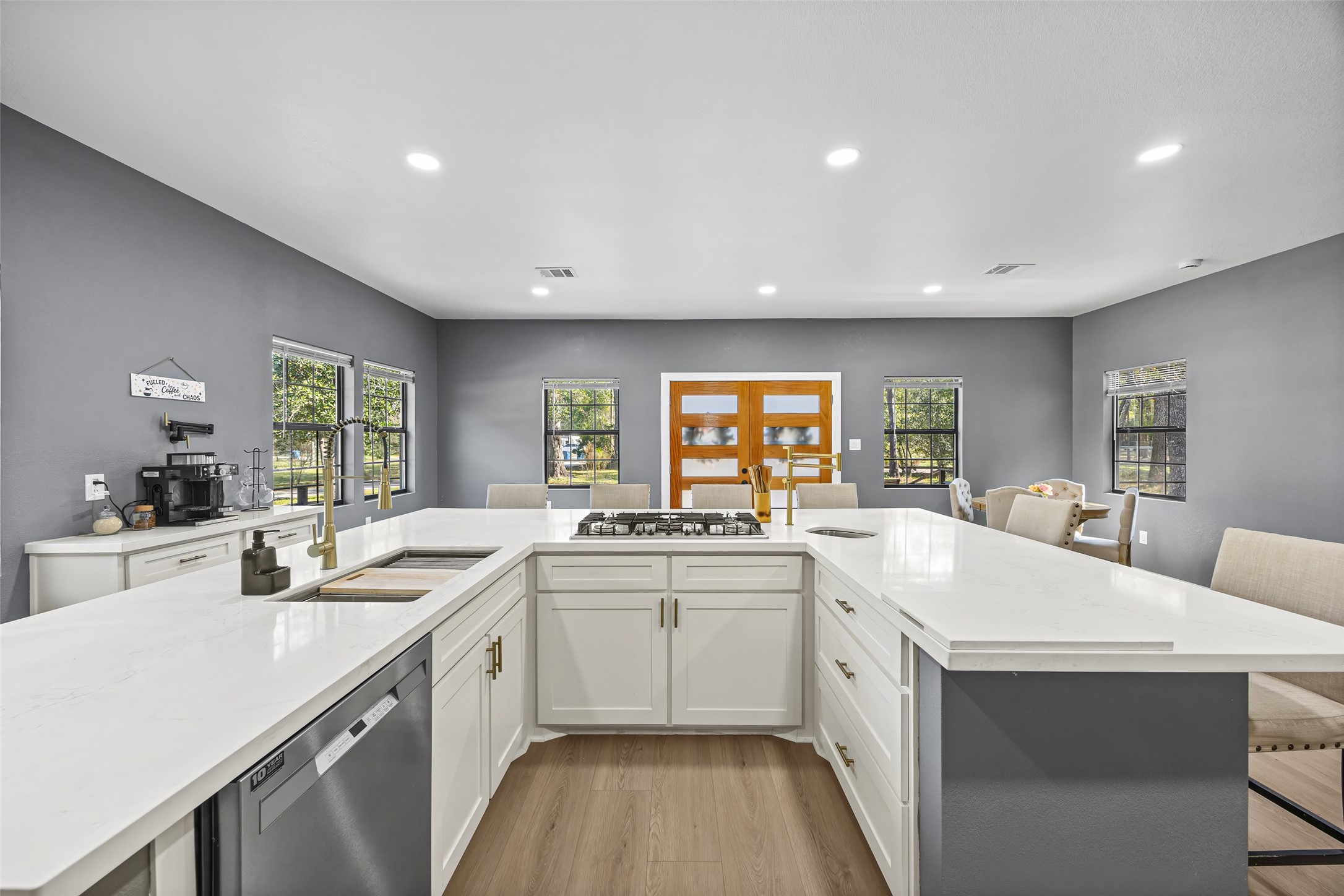 15080 Usher Lane Splendora, TX 77372 - Photo 19 of 44 Modern kitchen with sleek white cabinetry, large central island with a cooktop, and stainless steel appliances. The open layout features ample natural light from multiple windows, complemented by neutral gray walls and warm wood flooring.