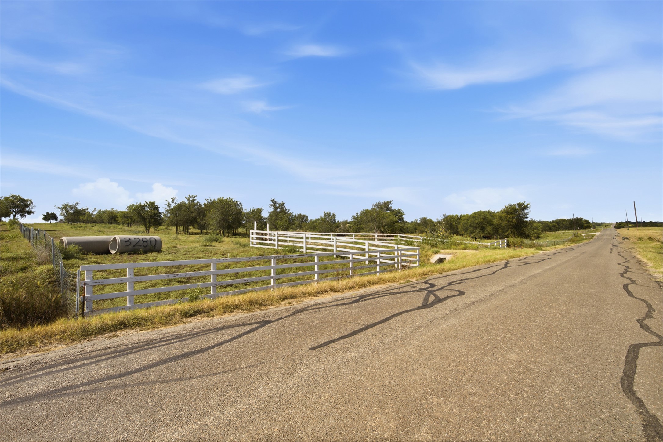 3280 County Road 466 Elgin, TX 78621 - Photo 10 of 13 An asphalt road meanders through a rural landscape, bordered by a classic white fence and scattered trees.