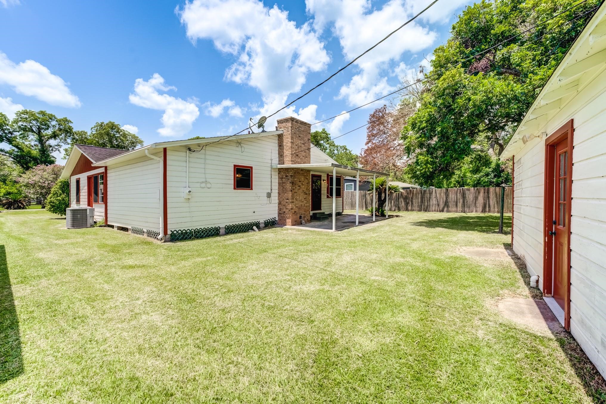 305 Yaupon Street Sweeny, TX 77480 - Photo 3 of 22 This photo shows a charming single-story home with a spacious backyard. It features a covered patio area, a brick chimney, and is surrounded by a wooden fence. The exterior is a combination of white siding and red trim, offering a cozy and inviting atmosphere.