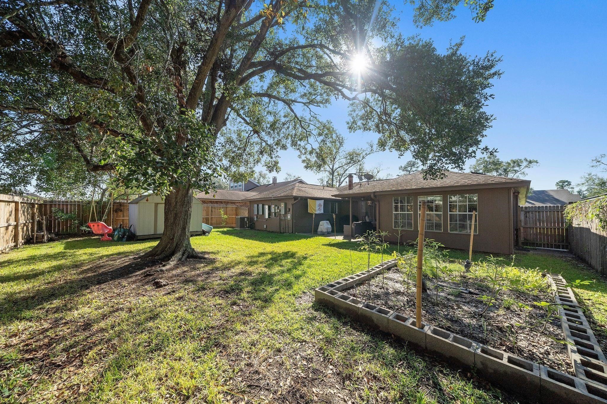 438 Vane Way Crosby, TX 77532 - Photo 16 of 21 This photo showcases a spacious backyard with a large tree providing shade, a garden bed, and a storage shed. The yard is fenced, offering privacy, and there's a patio area connected to the house, perfect for outdoor activities.