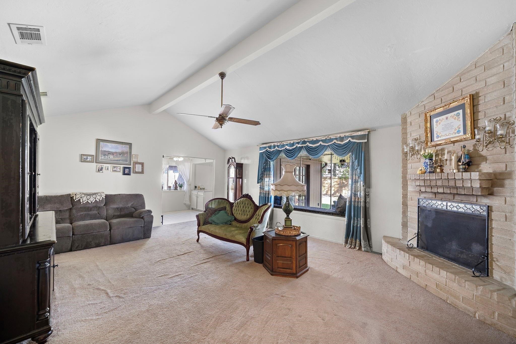 438 Vane Way Crosby, TX 77532 - Photo 2 of 21 This spacious living room features a vaulted ceiling, a cozy brick fireplace, and a large window with elegant drapes. It includes a ceiling fan, comfortable seating, and neutral carpeting, creating a warm and inviting atmosphere.
