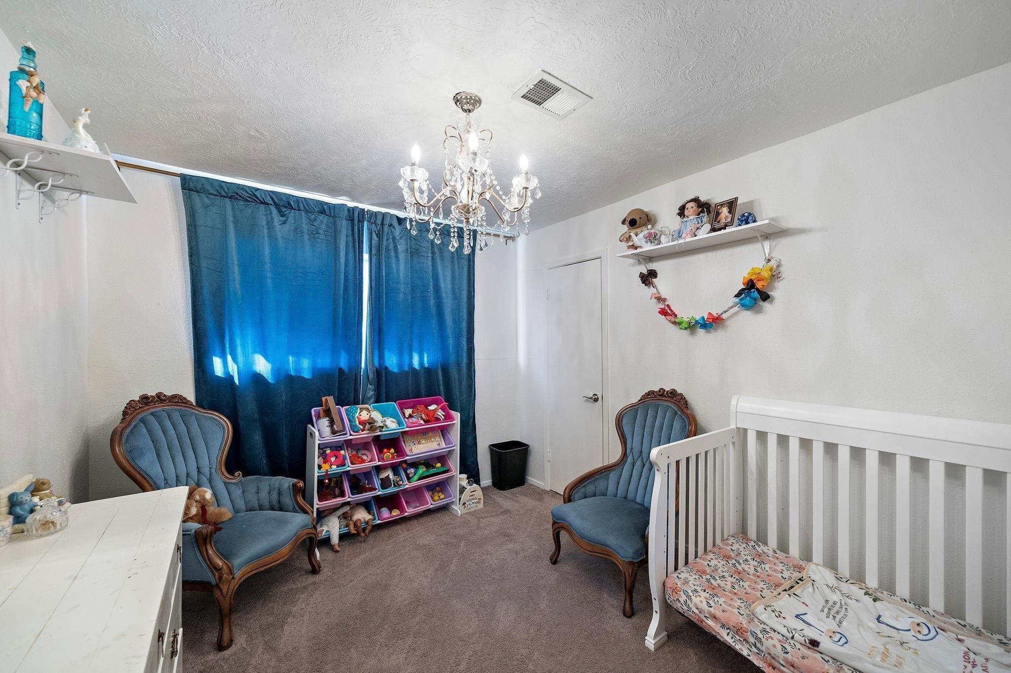 438 Vane Way Crosby, TX 77532 - Photo 7 of 21 This cozy nursery features a charming chandelier, soft carpet, and elegant vintage-style chairs. It includes a white crib and toy organizer, making it ideal for a child's room. The room is well-lit with a window covered by dark drapes, adding to its serene ambiance.