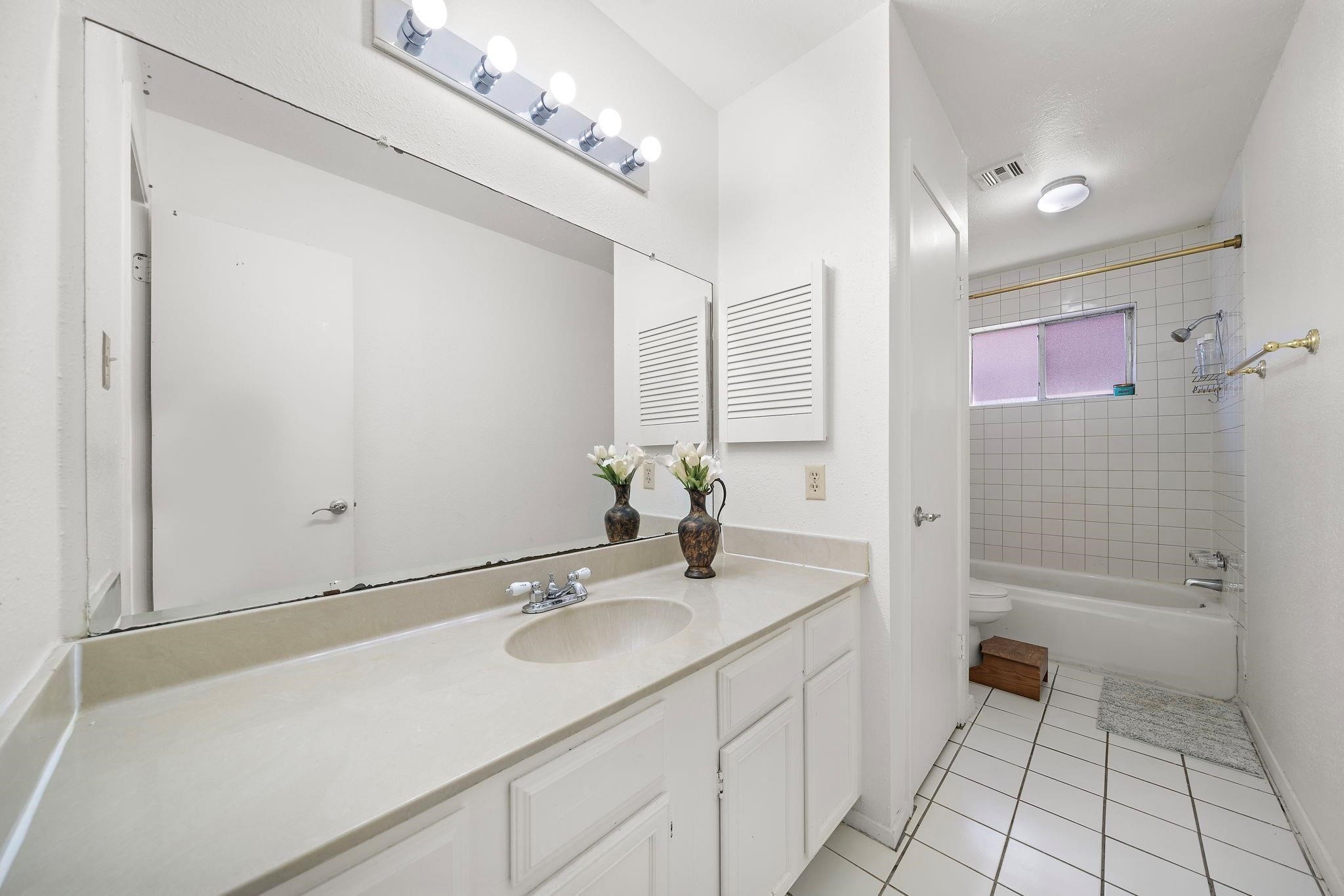 438 Vane Way Crosby, TX 77532 - Photo 8 of 21 This bathroom features a clean, bright design with a large mirror above a spacious countertop. It includes a bathtub with a shower, white tile flooring, and a simple, fresh aesthetic.