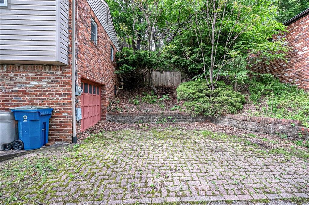 A spacious, brick-paver parking pad directly outside the garage, framed by sturdy stone retaining walls and mature trees for added privacy.