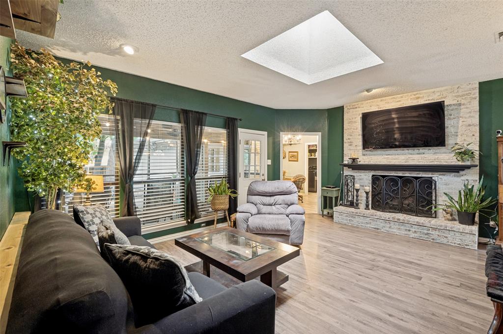 1006 Ridgeview Carrollton, TX 75007 - Photo 7 of 27 Living area featuring wood-finish flooring, a white brick fireplace with a black mantel, a large window offering natural light, and a prominent ceiling skylight