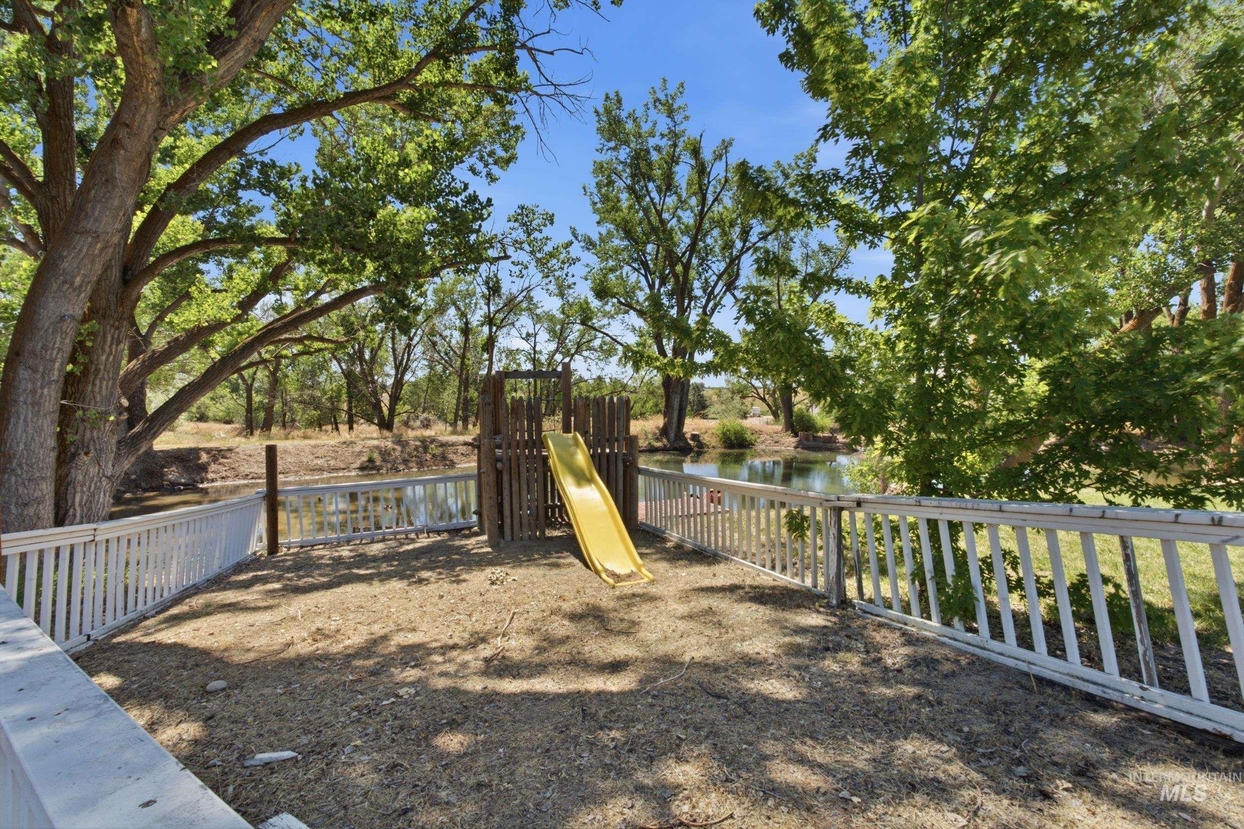 7781 Redmon Road Caldwell, ID 83607 - Photo 7 of 50 Fenced outdoor area featuring a wooden play structure with a yellow slide, mature trees, and a water feature beyond the white picket fence