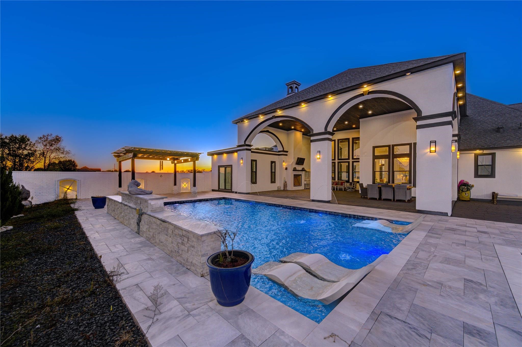 4821 Walters Road Richmond, TX 77469 - Photo 46 of 50 This stunning heated luxury pool creates a true resort-style experience with sleek modern design, expansive lounging areas, and breathtaking sunset views. Featuring elegant marble-style decking, in-water chaise loungers, ambient accent lighting, and a striking stone water feature, the space was thoughtfully designed for both relaxation and entertaining. A covered pergola lounge adds the perfect setting for evening gatherings, while the oversized pool offers a seamless blend of comfort & privacy.
