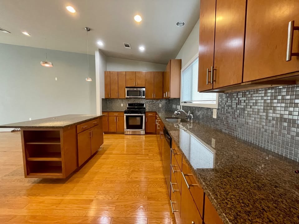 Kitchen featuring open shelves, brown cabinetry, dark stone counters, appliances with stainless steel finishes, and lofted ceiling