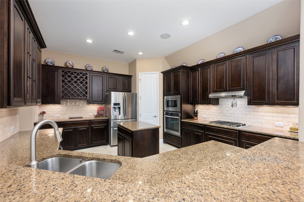 399 Whispering Wind Way Austin, TX 78737 - Photo 17 of 40 Kitchen with appliances with stainless steel finishes, under cabinet range hood, light stone counters, decorative backsplash, and dark brown cabinetry