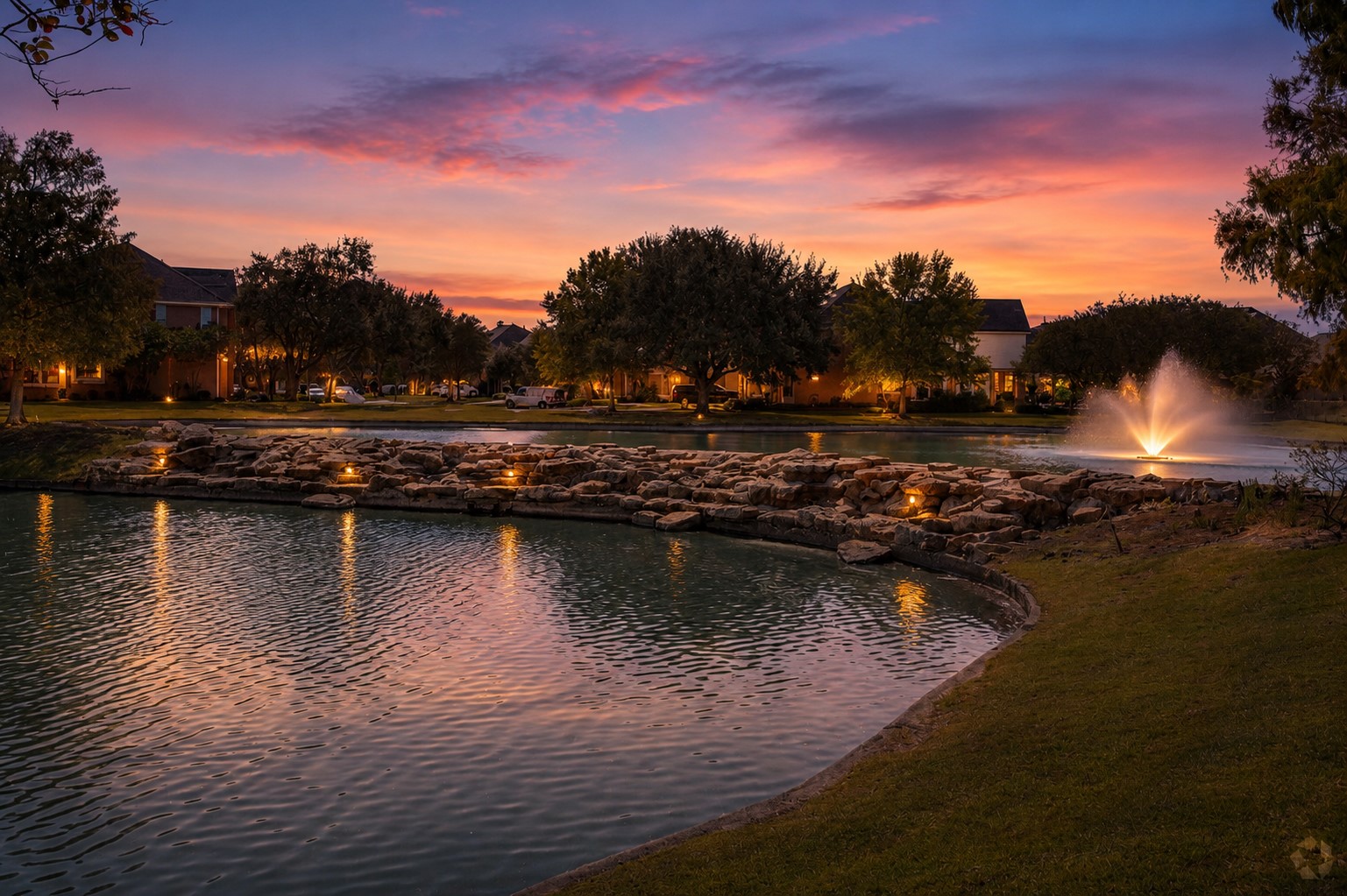 3118 Valley Court Manvel, TX 77578 - Photo 38 of 38 Neighborhood walk at sunset, featuring a tranquil pond with a rock-lined edge and an elegant fountain. The soft lighting highlights the surrounding trees and houses, creating a peaceful and inviting atmosphere.