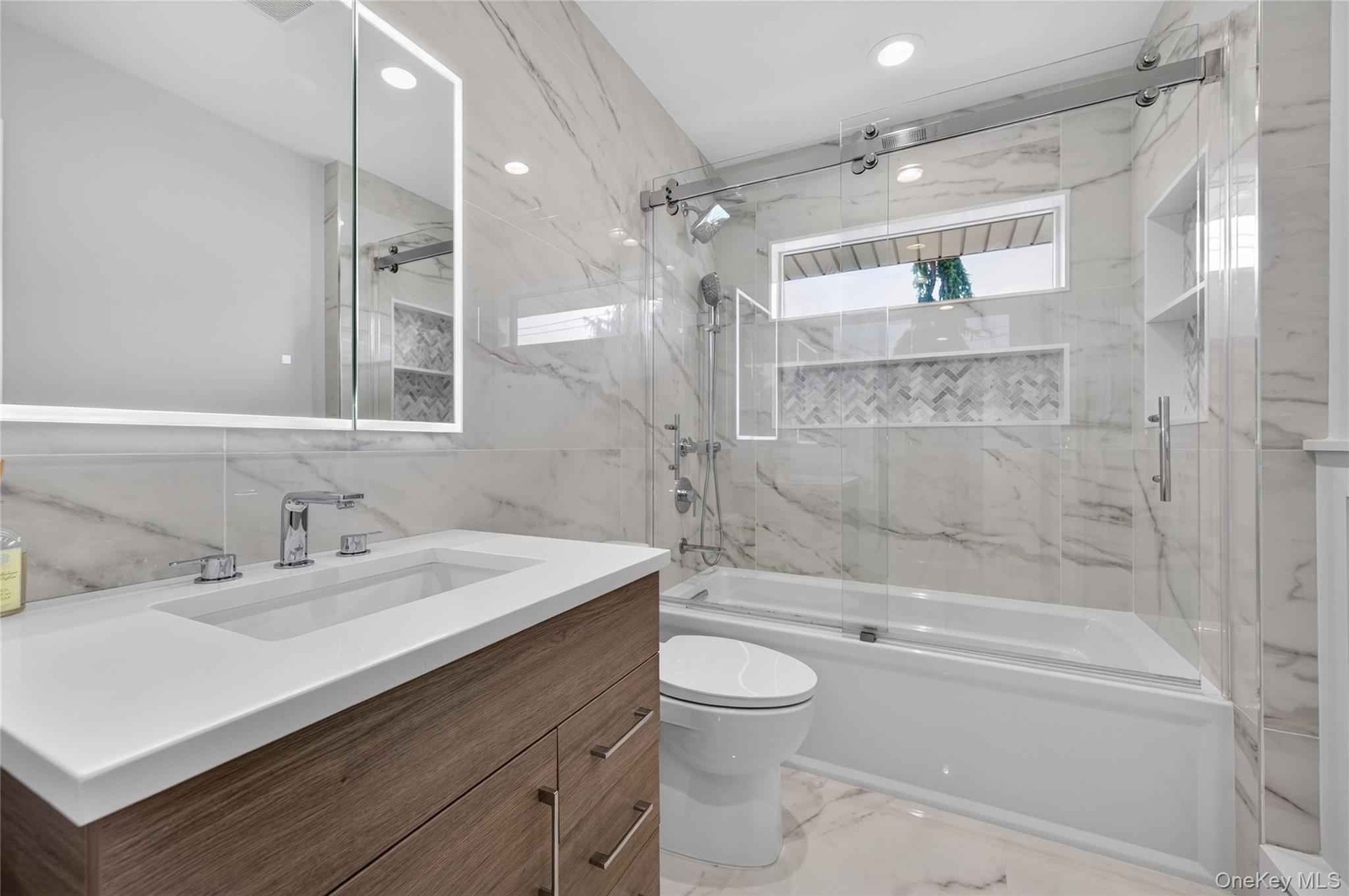11 Sunbeam Road Syosset, NY 11791 - Photo 16 of 28 Hall bathroom featuring a wood-finish vanity with an integrated sink and chrome faucet, an illuminated mirror, a tub with a sliding glass door enclosure with built-in niche