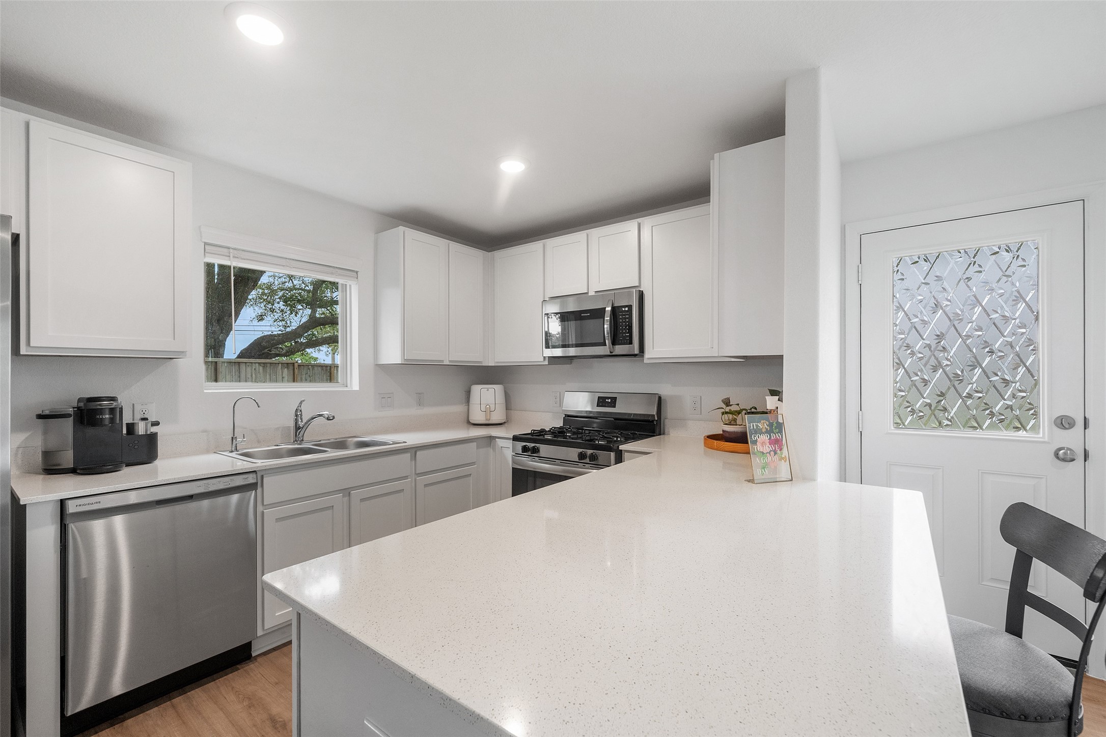 234 Bennett Street Angleton, TX 77515 - Photo 9 of 33 Modern kitchen with sleek white cabinetry, stainless steel appliances, and a large quartz countertop. Features a window with a view and a door with decorative glass, offering a bright and inviting space.