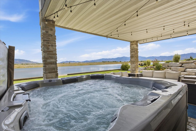 21 Pier Place Tooele, UT 84074 - Photo 94 of 112 Bubbling hot tub situated under a covered patio with string lighting, stone-clad pillars, and a serene lake backdrop