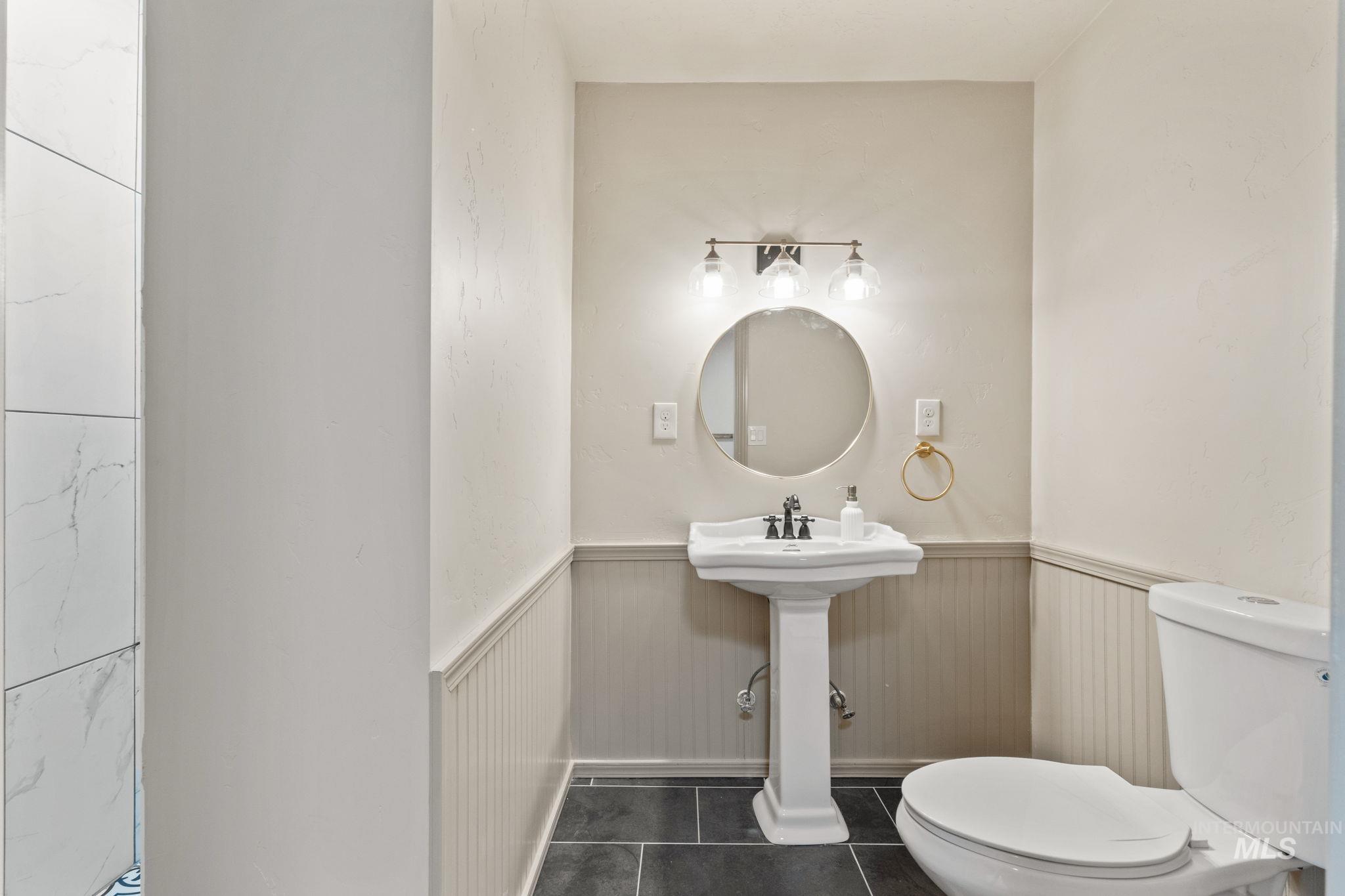 424 West Main Street Weiser, ID 83672 - Photo 12 of 30 Pedestal sink with a circular mirror, three-light vanity fixture, beadboard wainscoting, and dark tile flooring