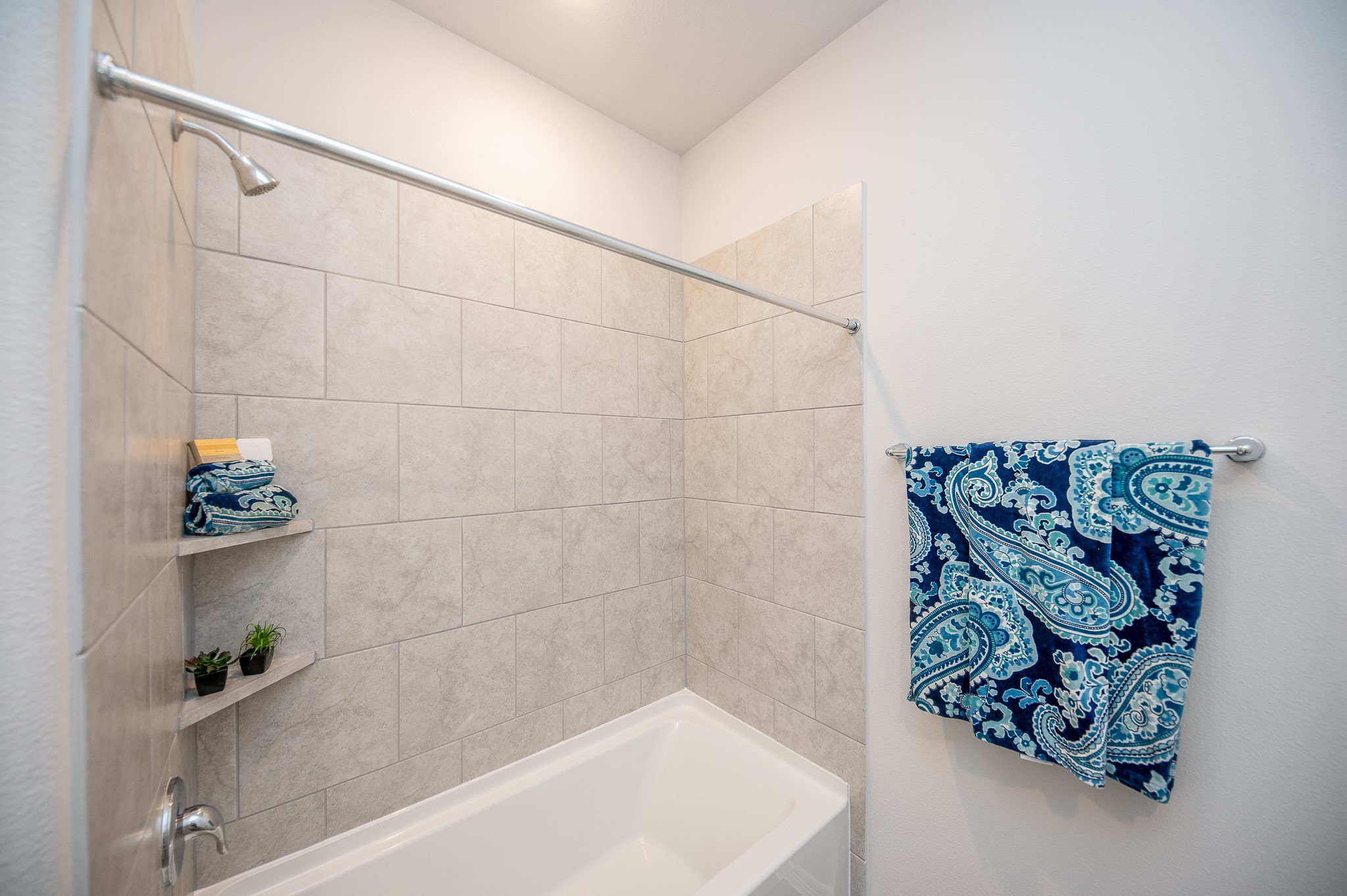 23302 Spring Genesis Lane Katy, TX 77493 - Photo 30 of 32 This bathroom features a clean, modern design with light beige tiled walls and a white bathtub. It includes built-in shelving for storage and a stylish blue patterned towel for a touch of color.