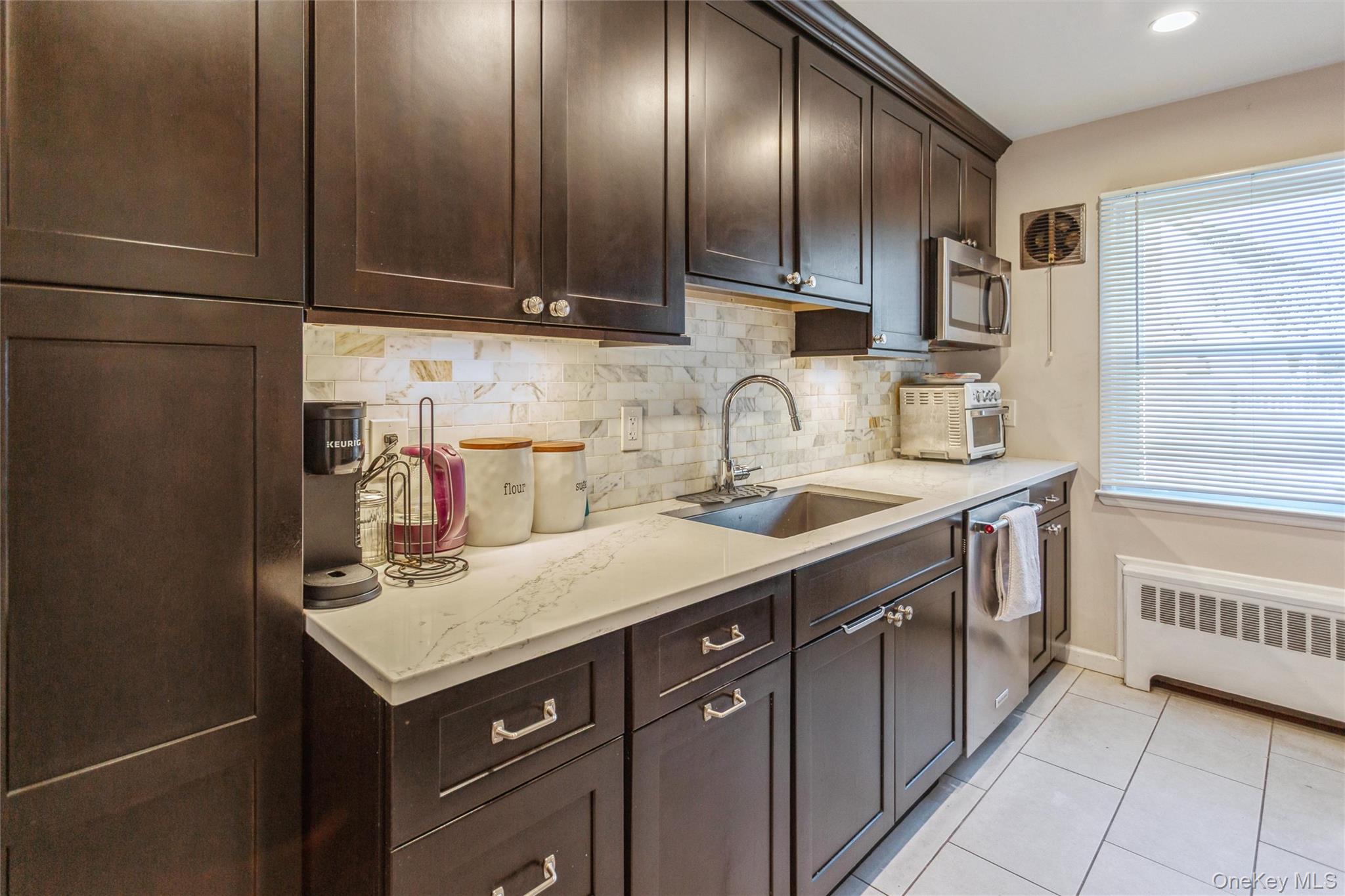 1531 Merrick Avenue Merrick, NY 11566 - Photo 13 of 26 Kitchen featuring dark brown cabinets, backsplash, light tile patterned flooring, radiator, and light stone countertops