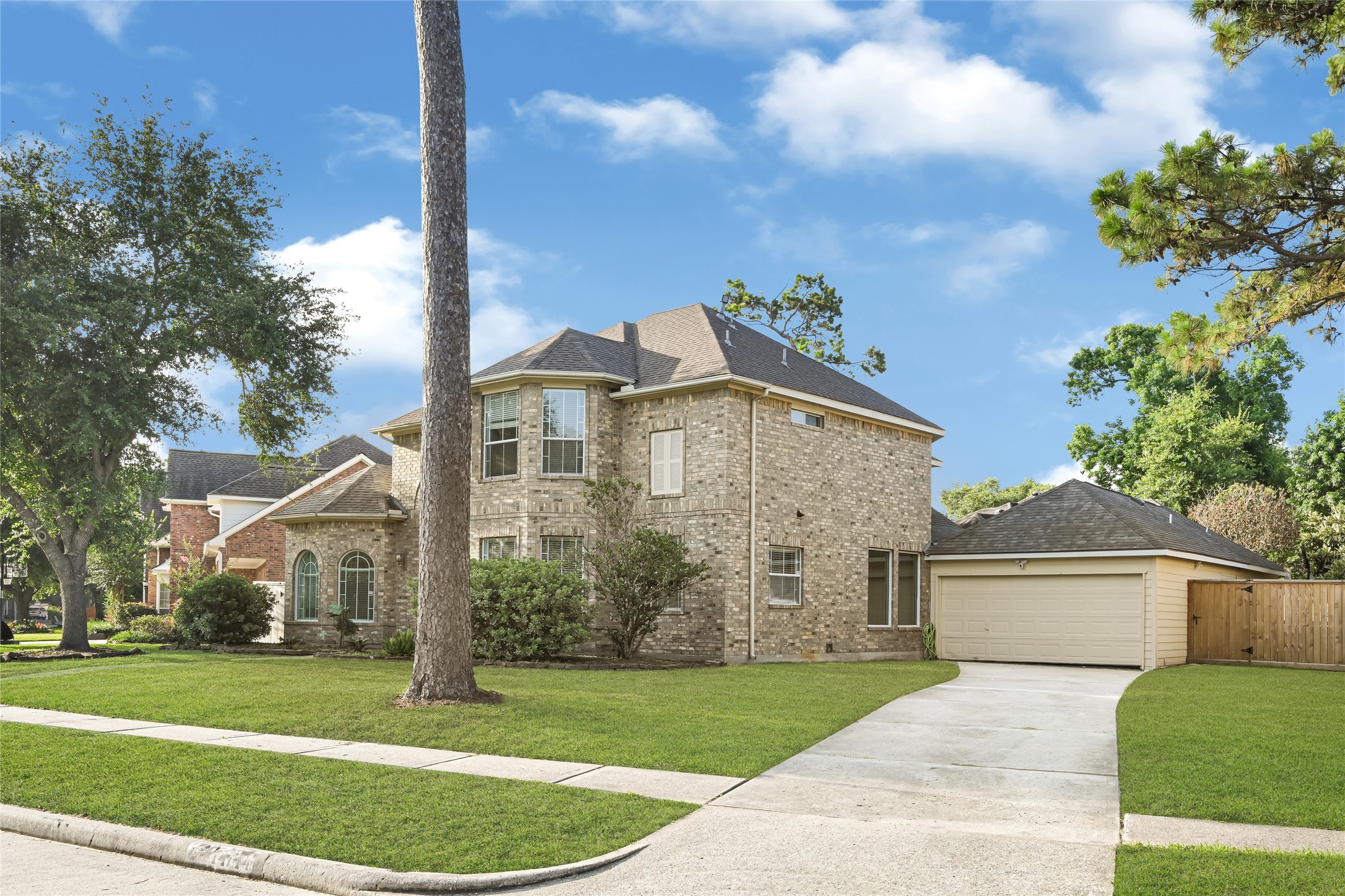 This is a charming two-story brick home with a well-maintained lawn and mature trees. It features a spacious driveway leading to a detached garage, and the neighborhood appears peaceful and green.