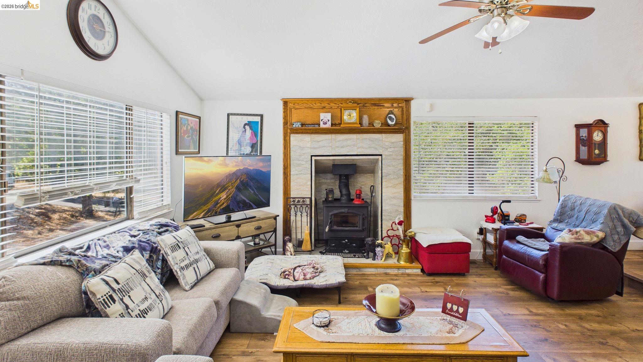 11858 Campo Seco Road Sonora, CA 95370 - Photo 34 of 45 Living area featuring vaulted ceilings, wood-finish flooring, a wood-burning stove with a stone surround and wood mantel, large windows with horizontal blinds, and a ceiling fan