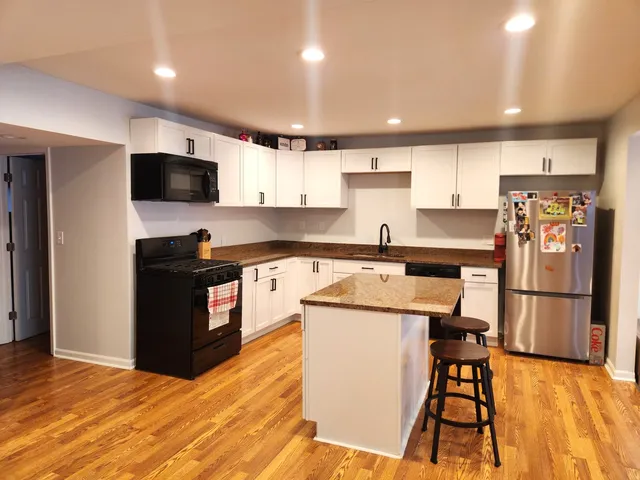a kitchen with stainless steel appliances kitchen island granite countertop a refrigerator a stove top oven a sink and white cabinets with wooden floor