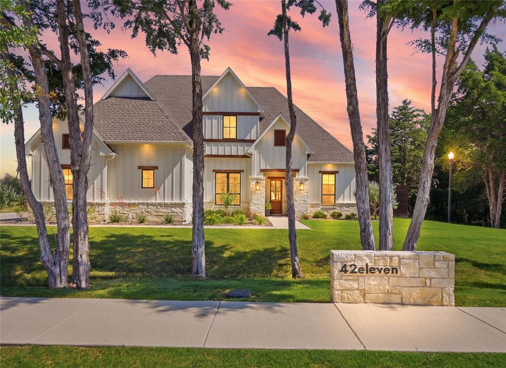 Modern farmhouse exterior featuring board and batten siding, stone veneer accents, a dark shingled roof, and an illuminated stone address monument