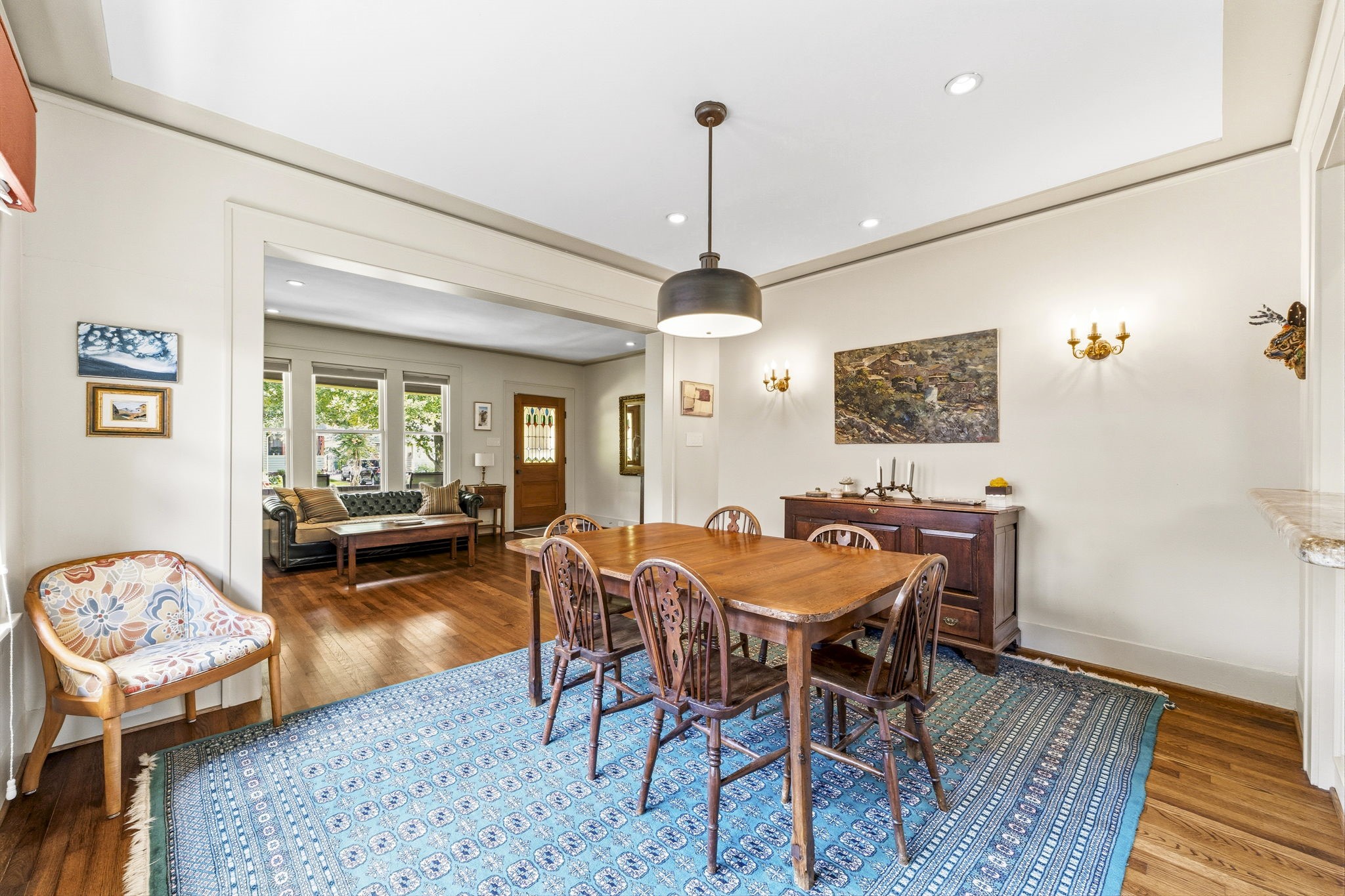 1521 Kipling Street Houston, TX 77006 - Photo 13 of 35 The photo showcases a bright, inviting dining area with a wooden table and chairs on a patterned blue rug. The room features hardwood floors, a modern light fixture, and wall sconces. It opens to a cozy living space with large windows, allowing natural light to fill the area, highlighting the classic and elegant decor.