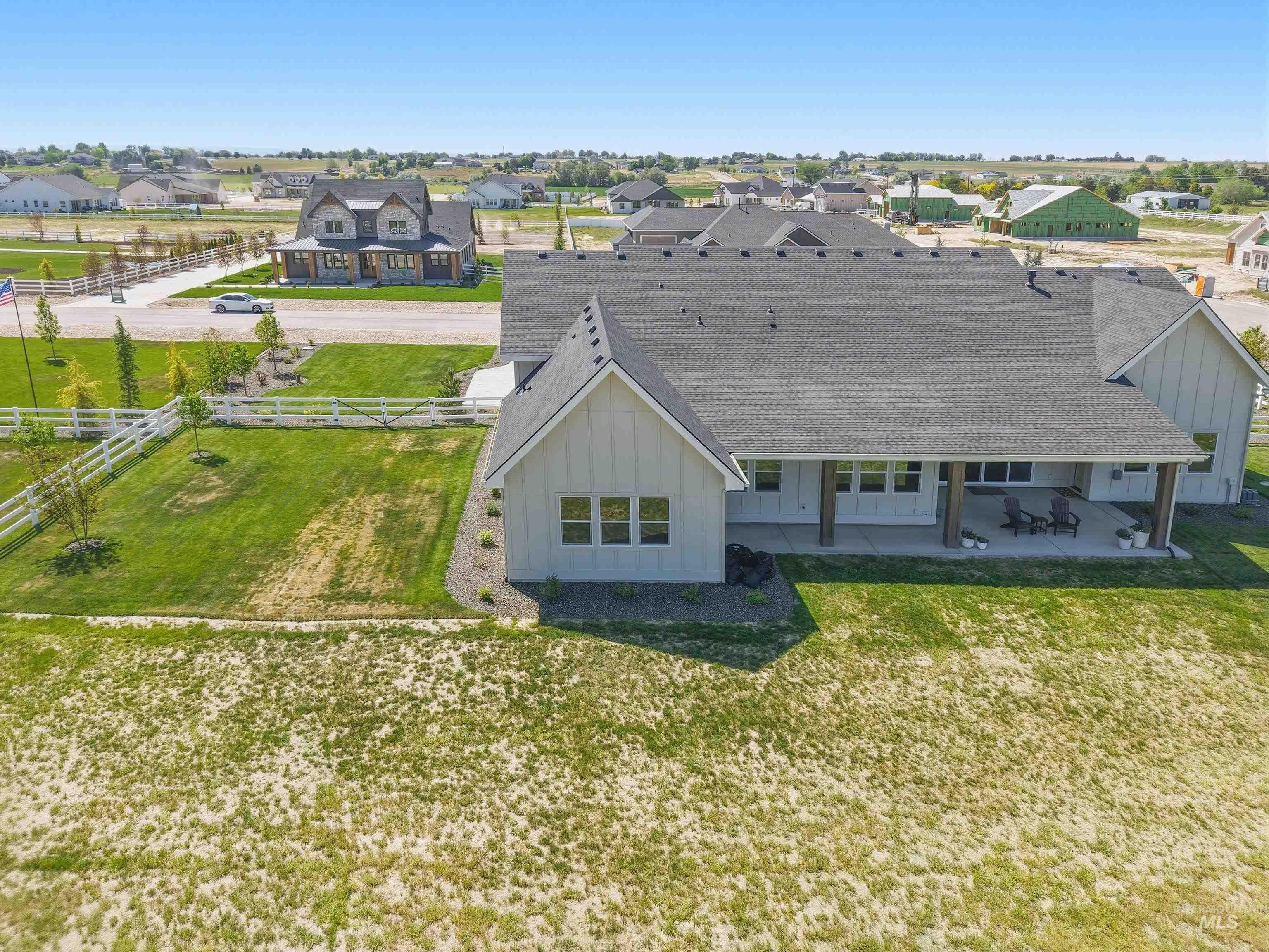 24990 Belgian Way Middleton, ID 83644 - Photo 50 of 50 Modern farmhouse-style residence featuring a dark shingle roof, light-colored board and batten siding, multiple windows, an expansive covered patio with wooden support beams, and a large grassy yard with a white picket fence