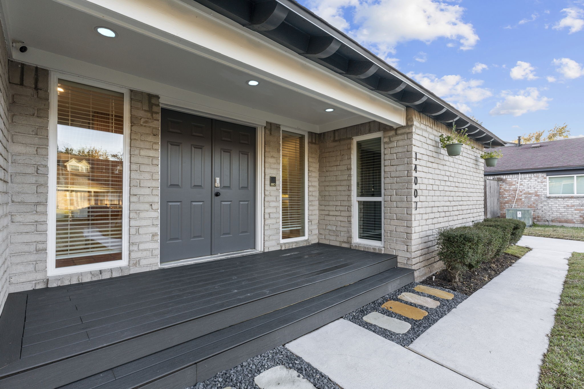 This home features a welcoming front entrance with a modern double door, sleek dark decking, and large windows. The exterior is crafted with light brick and complemented by a neat pathway and tidy landscaping.