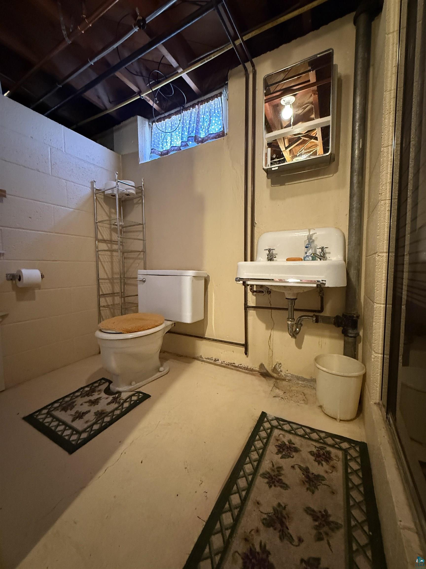 607 3rd Street Northeast Chisholm, MN 55719 - Photo 27 of 32 Functional bathroom featuring exposed ceiling joists, a wall-mounted sink with a mirrored medicine cabinet, a toilet, and a window with curtain