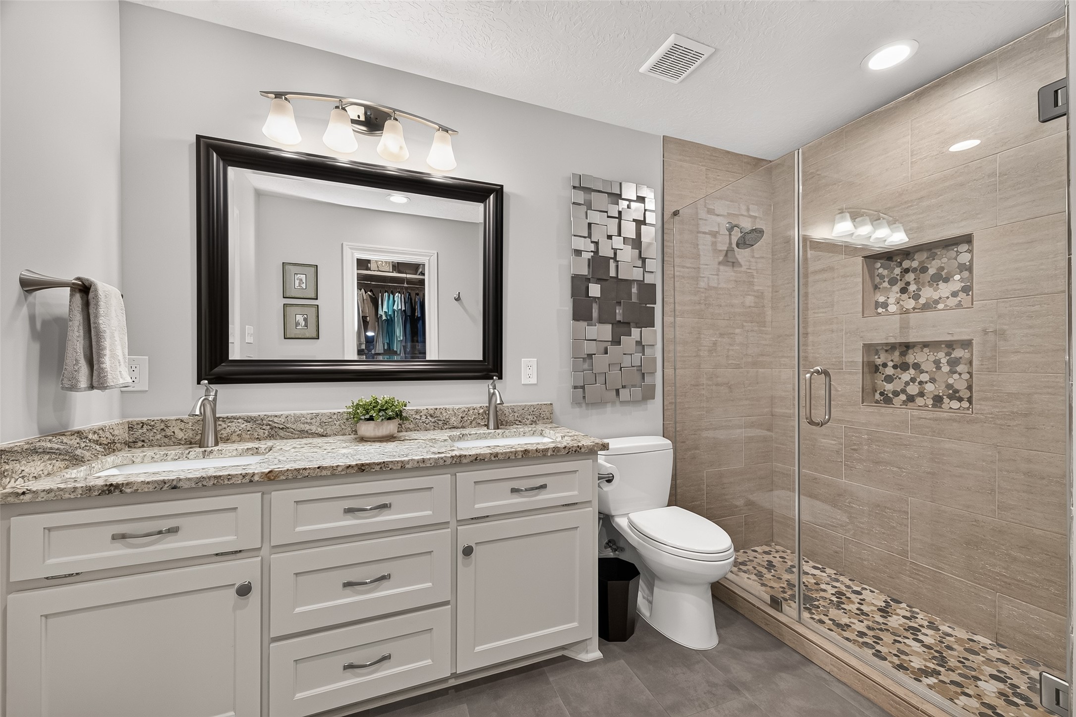 24719 Wilderness Road Spring, TX 77380 - Photo 32 of 42 This bathroom features a modern design with a double-sink vanity, granite countertops, and a large framed mirror. It includes a spacious glass-enclosed shower with textured tile and built-in shelving. The room is well-lit with contemporary fixtures, creating a bright and inviting atmosphere.
