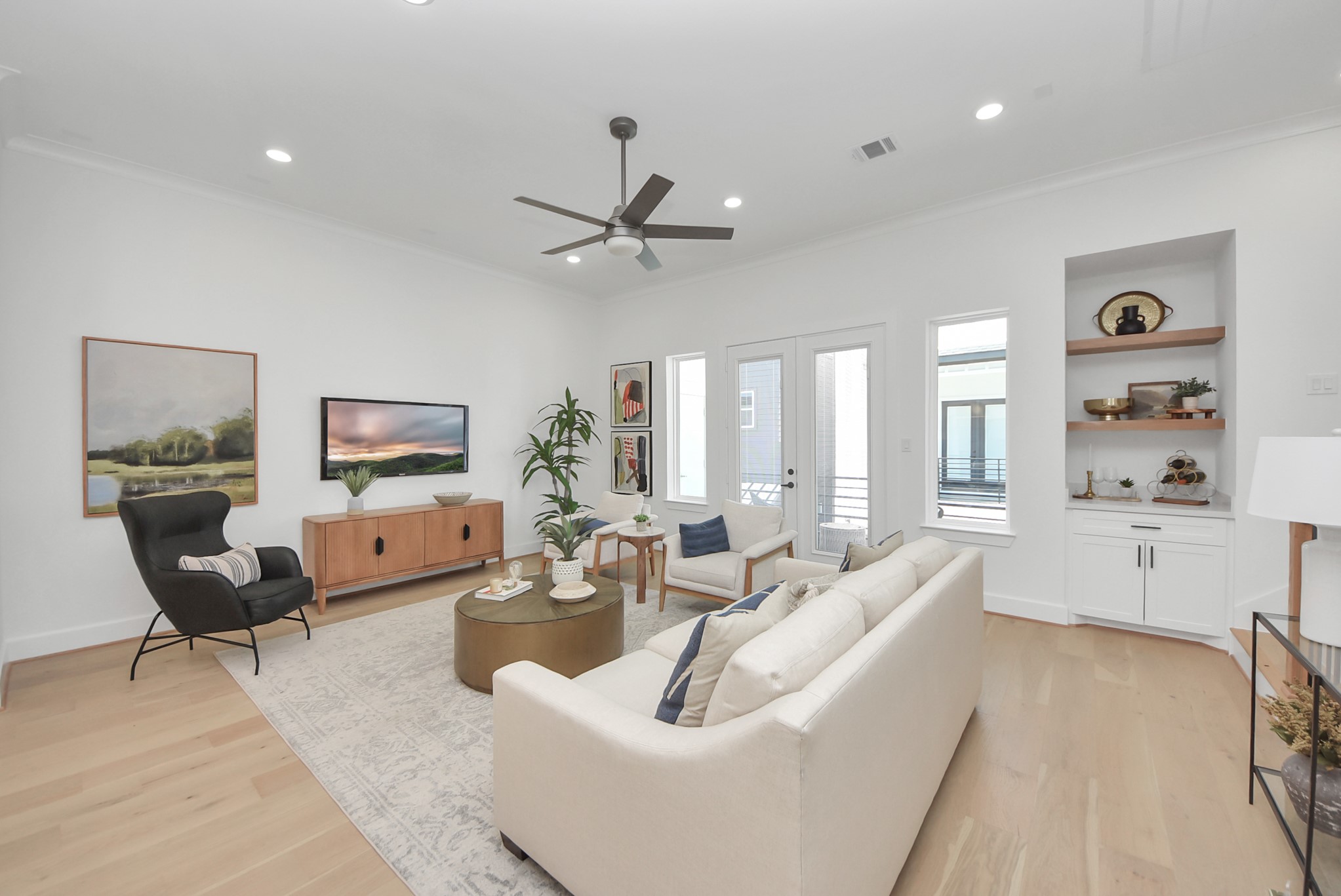 927 Fisher Street, Unit B Houston, TX 77018 - Photo 14 of 47 Step into this bright and airy living room, where modern decor and light wood flooring create an inviting atmosphere. A cozy sofa and armchair complement the sleek ceiling fan, while built-in shelves, large windows, and doors to the outdoor area seamlessly blend functionality with style—perfect for both relaxation and entertaining.