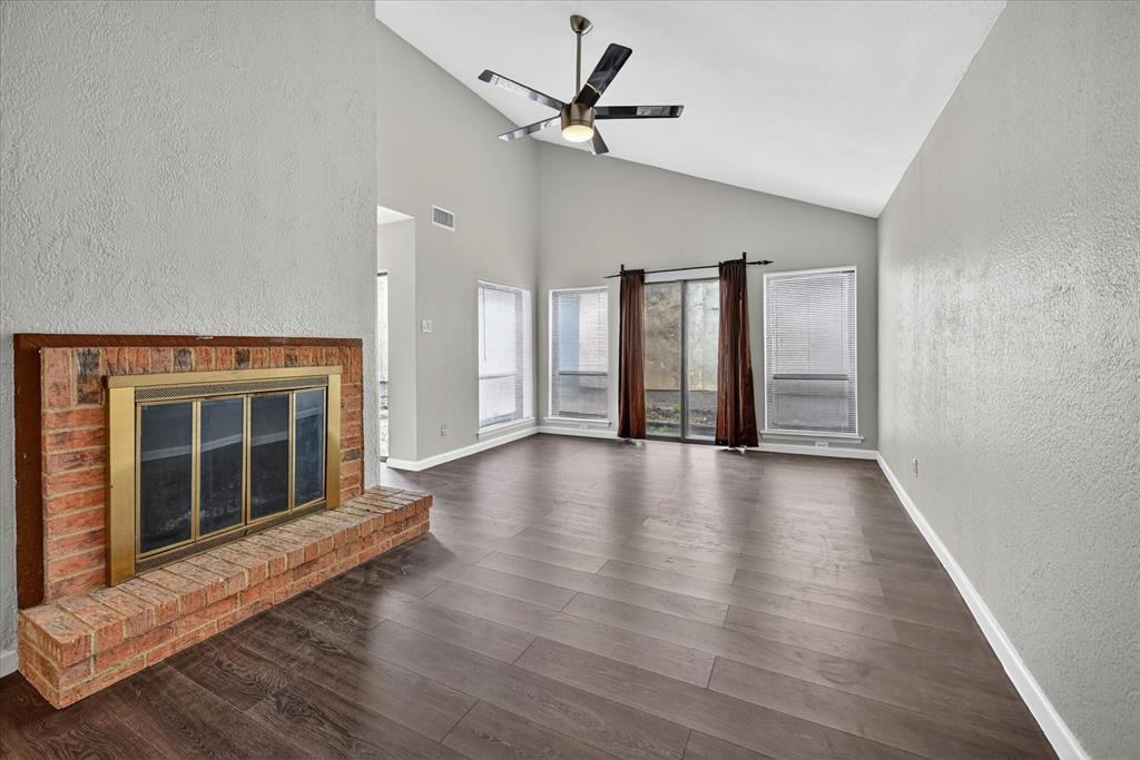 9809 Matchpoint Place Dallas, TX 75243 - Photo 4 of 21 Vaulted ceiling living area featuring a brick fireplace with a brass firebox, dark wood-finish flooring, and multiple large windows with vertical blinds