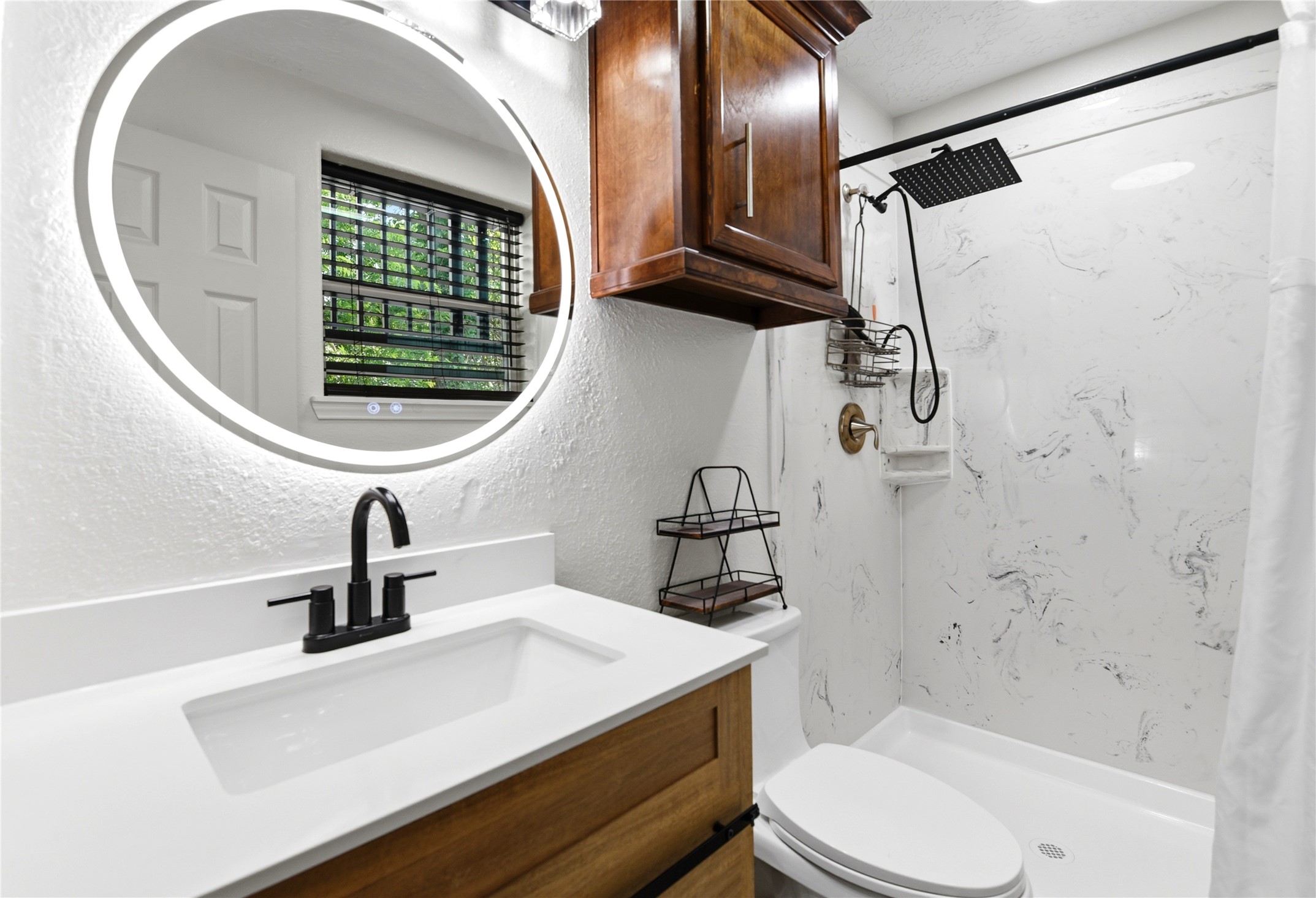 9403 Spaulding Street Houston, TX 77016 - Photo 24 of 50 This bathroom features a modern design with a circular backlit mirror, a sleek black faucet, and a wood vanity. There's a walk-in shower with a rainfall showerhead and marble-style walls. Natural light streams through a window with blinds, complementing the cozy, elegant feel.