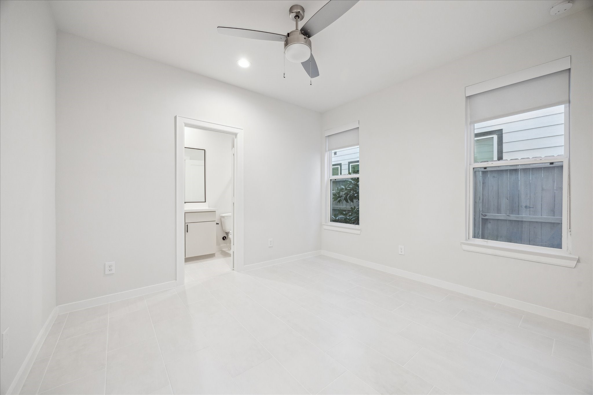1603 Patterson Street, Unit E Houston, TX 77007 - Photo 9 of 32 This photo showcases a bright, modern bedroom with light-colored flooring and walls. It features a ceiling fan, two windows with blinds, and an en-suite bathroom with a visible vanity and mirror, creating a clean and inviting space.