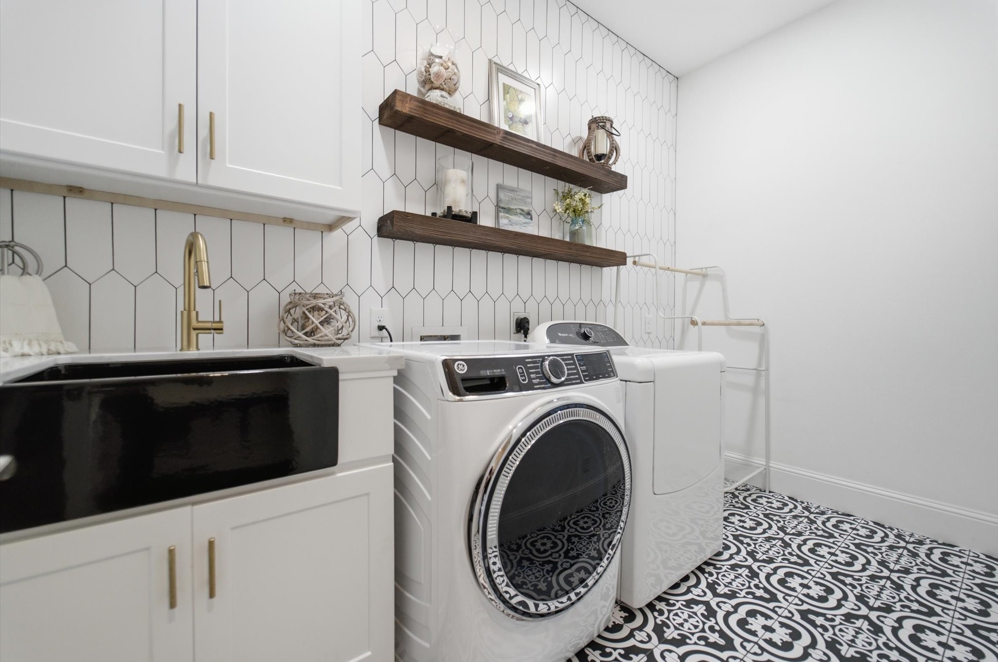 23722 Thortons Park Lane Spring, TX 77389 - Photo 17 of 48 This photo showcases a stylish laundry room with a modern washer and dryer, sleek black and white patterned tile flooring, white cabinetry, gold hardware, and a black sink. The hexagonal tile backsplash and wooden shelves add a touch of warmth and elegance.
